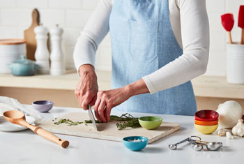 a woman chops alongside herbs prep bowls while wearing a blue kitchen apron