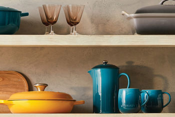 an assortment of colorful Le Creuset cookware on shelves including a french press and mugs